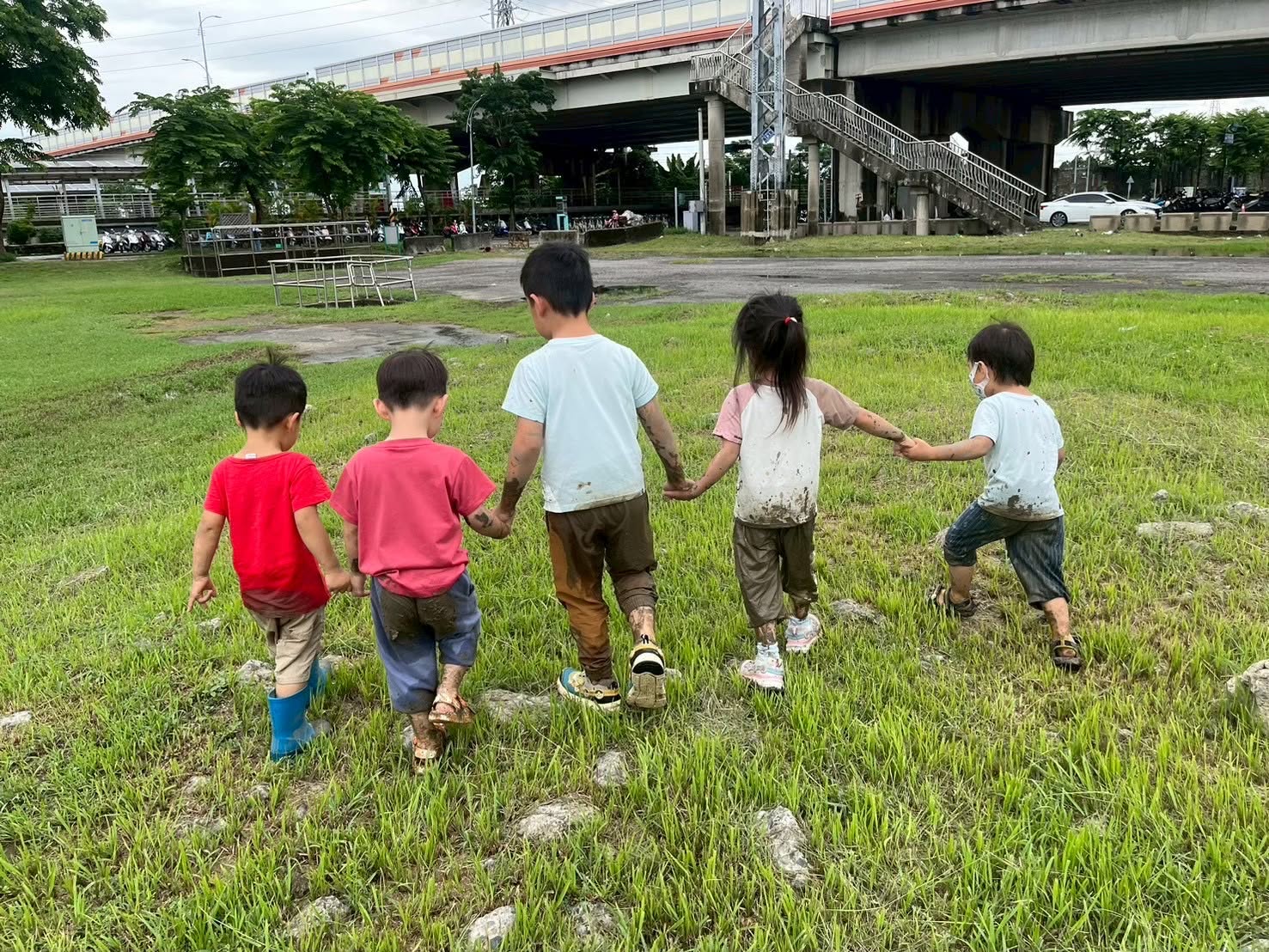 五位孩子手牽著手走過草地，衣服與鞋子上留著玩泥水的痕跡