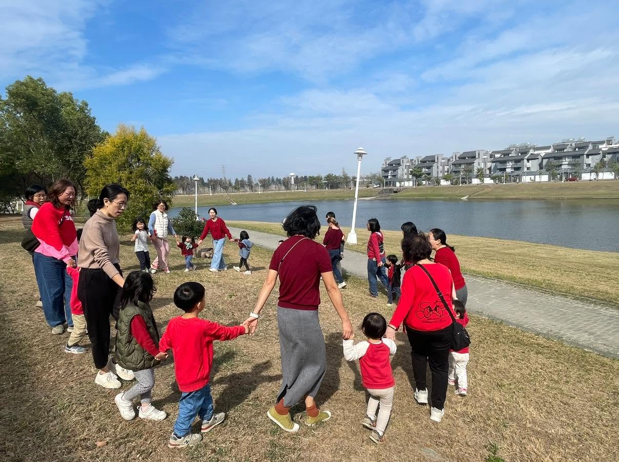 家長與孩子在水岸旁的草地上牽手圍成圈，一起參與戶外活動
